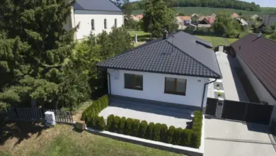 Bílý rodinný dům s tmavou taškovou střechou v přírodě.