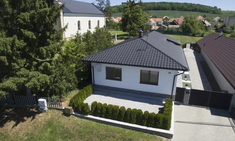Bílý rodinný dům s tmavou taškovou střechou v přírodě.