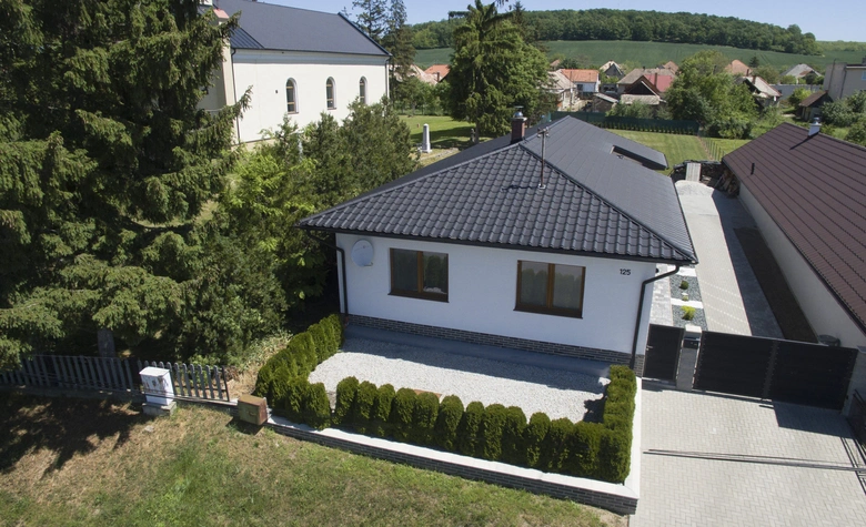 Bílý rodinný dům s tmavou taškovou střechou v přírodě.