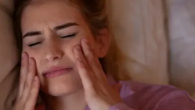 Žena leží v posteli s bolestí čelisti a drží si tváře.