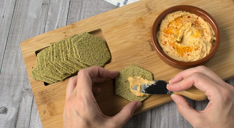 Ruce rozmazávají hummus na zelené sušenky vedle misky.