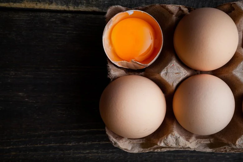 Čtyři vejce v kartonové krabici s rozbitou skořepinou ukazující žloutek.